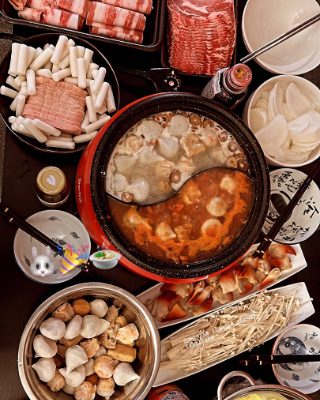 A hot pot spread — a split broth pot ringed by sliced meat, rice cakes, enoki mushrooms, tofu and cabbage.