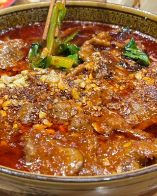 A wide bowl of Sichuan boiled beef in chilli oil, scattered with peppercorns and green chillies.
