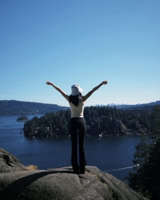 A figure standing on Quarry Rock above Deep Cove with both arms out, the inlet and forested islands below.