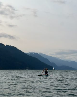 A small distant figure paddleboarding on wide gray water with layered mountains behind.