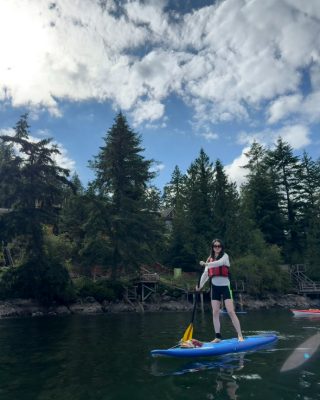 Standing upright on a blue paddleboard in a cove ringed by evergreens.